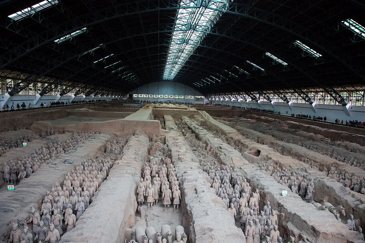 Pit 1 of the Terracotta Army: rows of clay warriors in earthen corridors separated by walls, under the metal roof of the hall, with visitors along the far edge.