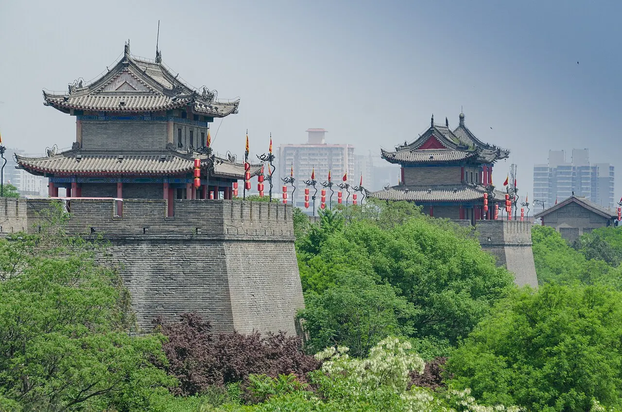 Two curved-roof guard pavilions on Xi'an's grey brick city wall, with red lanterns and flags on top, green trees below and tall buildings behind.