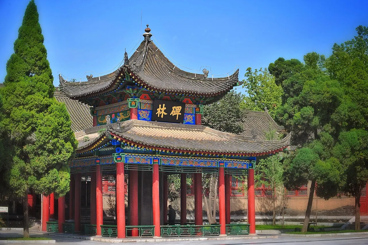 The entrance pavilion of the Forest of Stelae in Xi'an, with two stacked roofs, red columns and a plaque with gold characters, among cypresses and trees under a blue sky.
