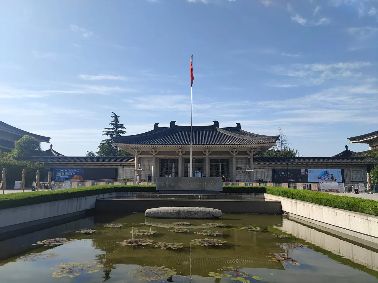 The front of the Shaanxi History Museum, a dark-roofed building in traditional style with a flagpole and red flag, behind a reflecting pool with water plants.