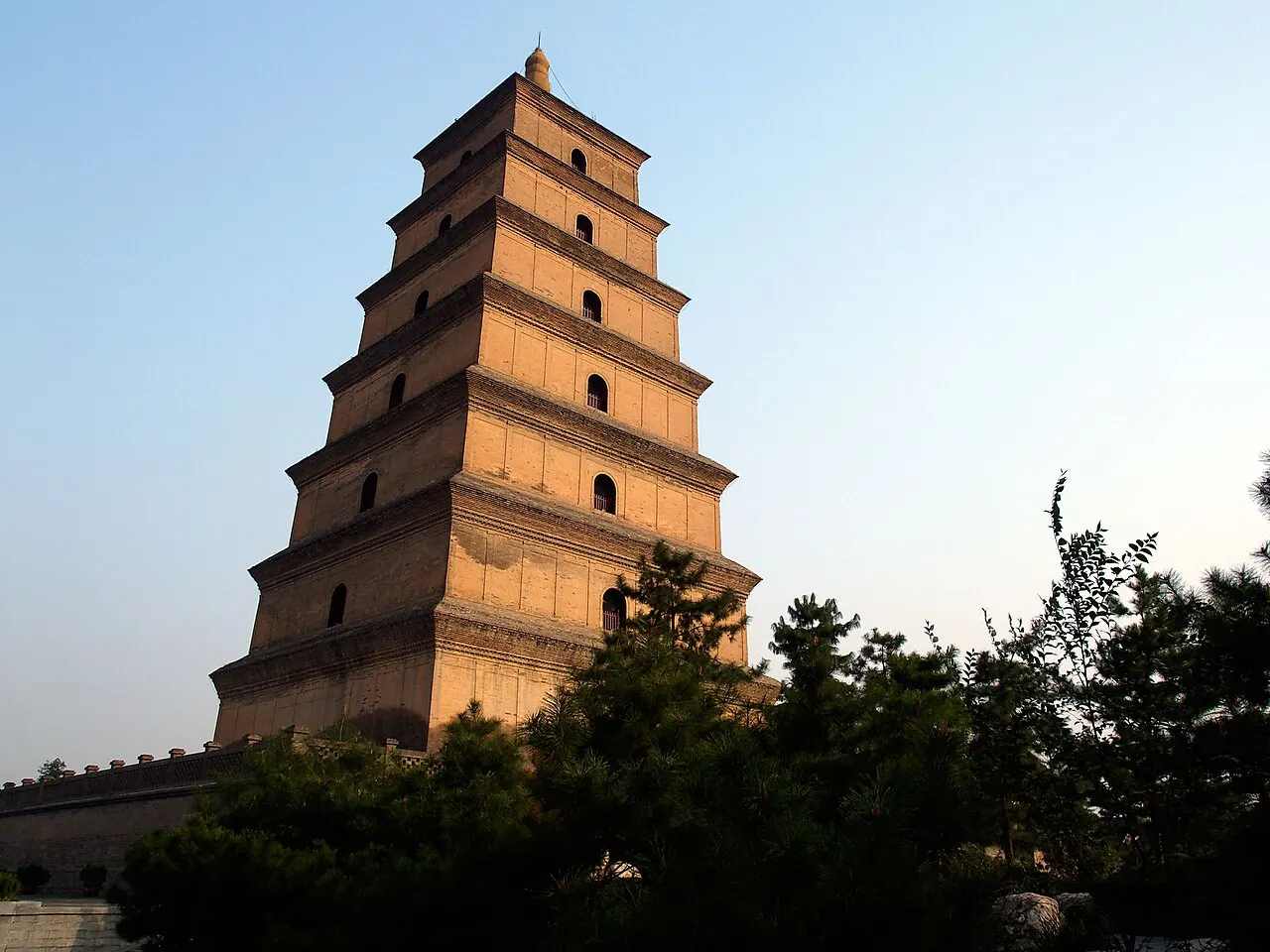The Big Wild Goose Pagoda, a square seven-storey brick tower with small arched windows, lit by warm light above dark pine trees.