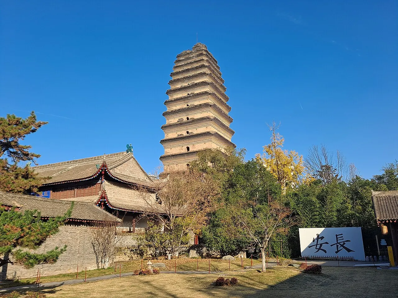 The Small Wild Goose Pagoda, a brick tower of many narrow eaves with a flat top, behind a curved-roof temple hall and autumn trees, under a blue sky.