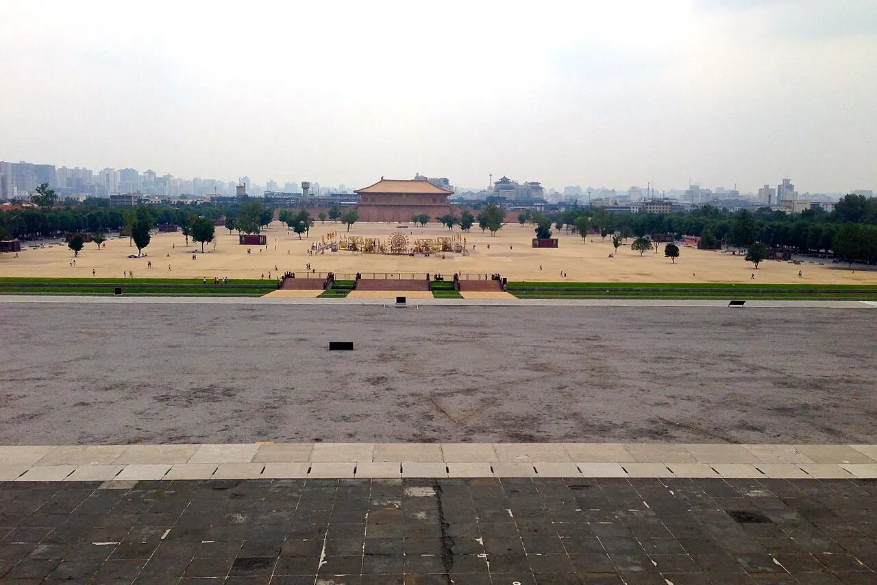 The view from the platform of a Daming Palace hall: an open ground of earth and lawn with scattered trees stretching to the rebuilt Danfeng Gate, with city buildings on the horizon.
