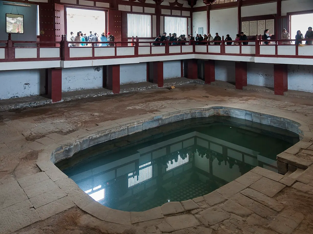 An excavated ancient stone bath at Huaqing Palace, filled with green water, inside a hall with a red wooden gallery where visitors look down.