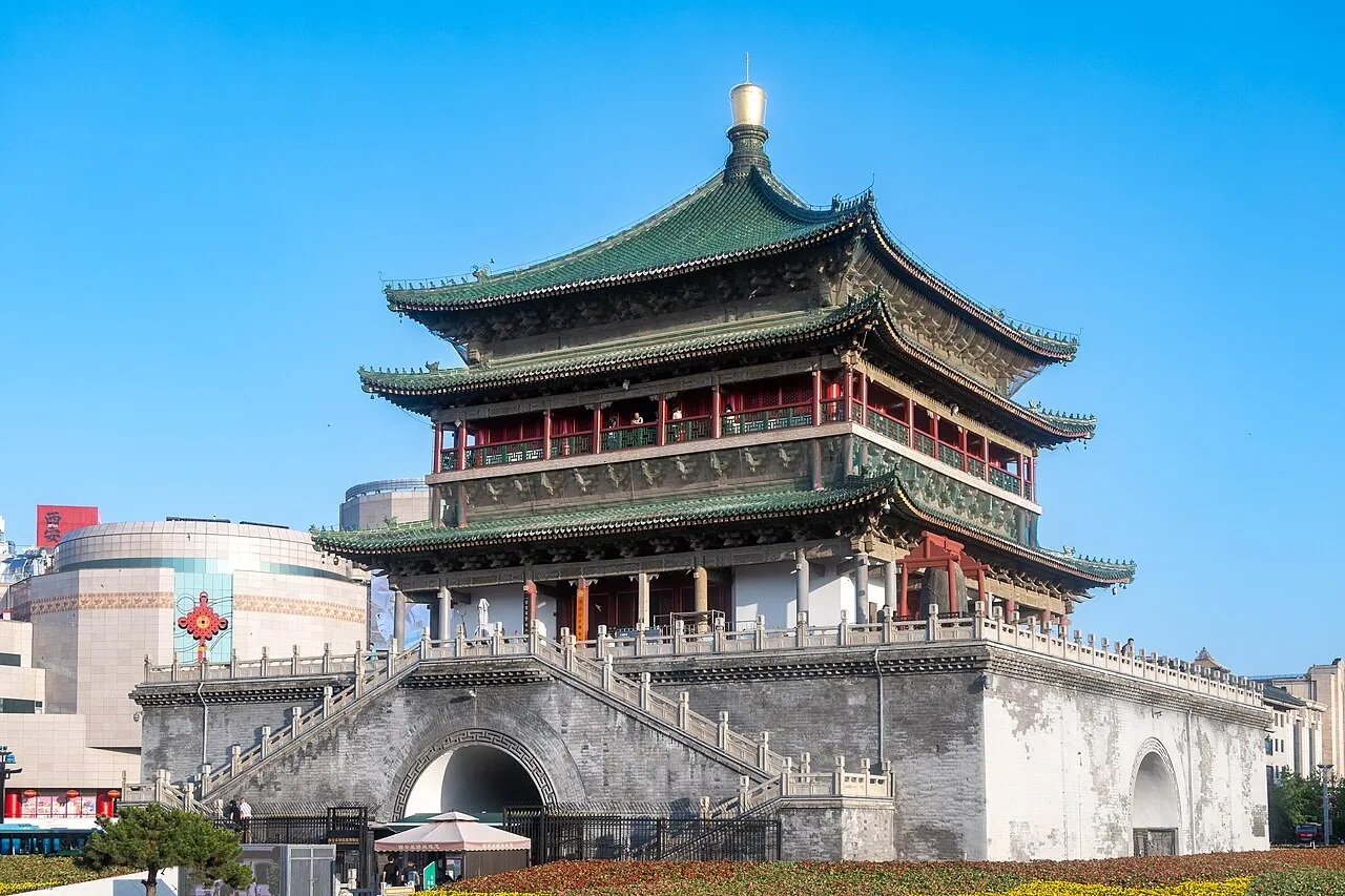 Xi'an Bell Tower, with three tiers of green-tiled eaves and red balconies on a square brick base with an archway and stairs, flower beds in front and a blue sky.