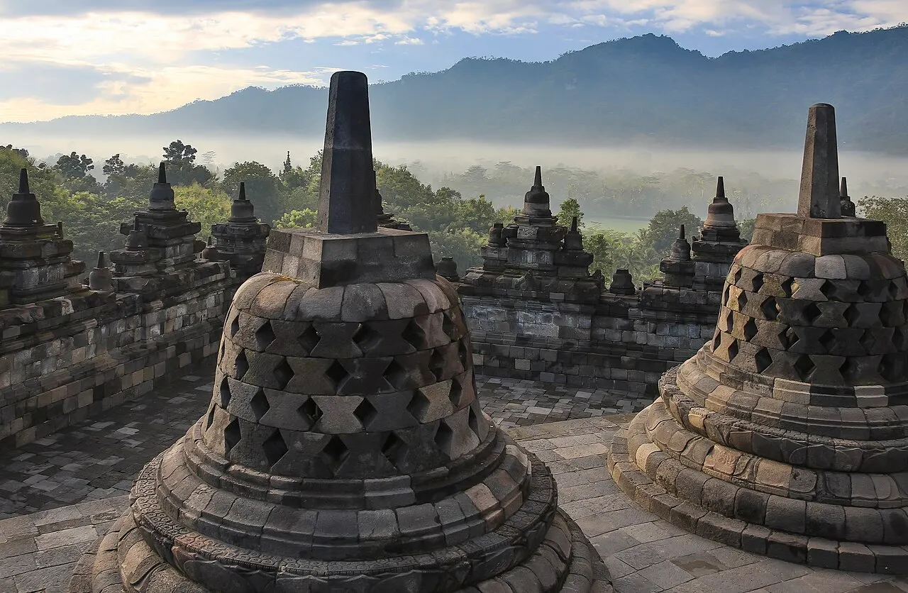 Duas estupas de pedra em forma de sino, com aberturas em losango, num terraço do templo, com névoa sobre a floresta e montanhas ao fundo.