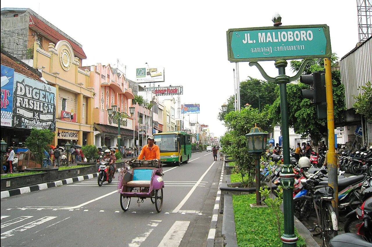 Rua comprida com lojas de fachadas coloridas, um becak e motos na pista, ônibus verde ao fundo e uma placa verde com o nome da rua num poste.