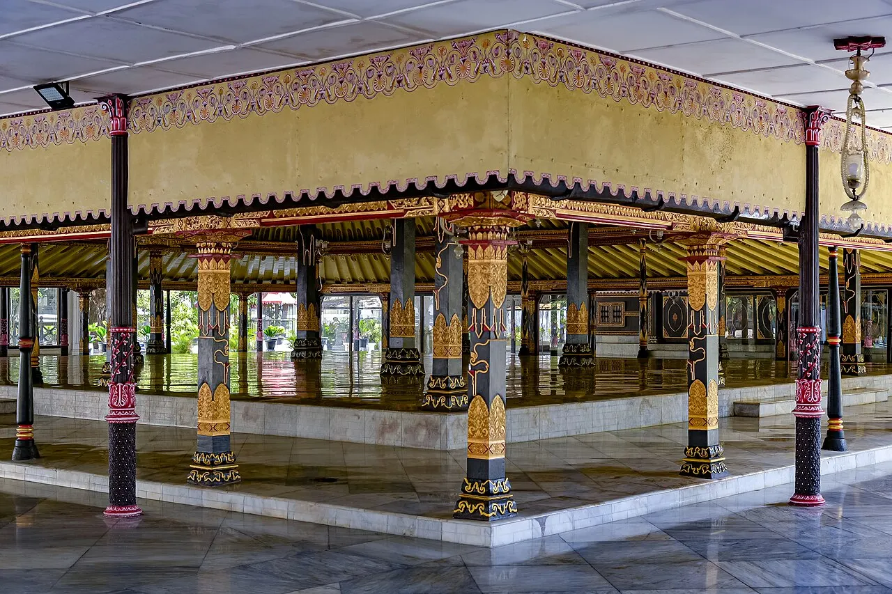Pavilhão aberto de piso de mármore, com colunas escuras decoradas em dourado e vermelho sob uma cornija amarela pintada com motivos florais.