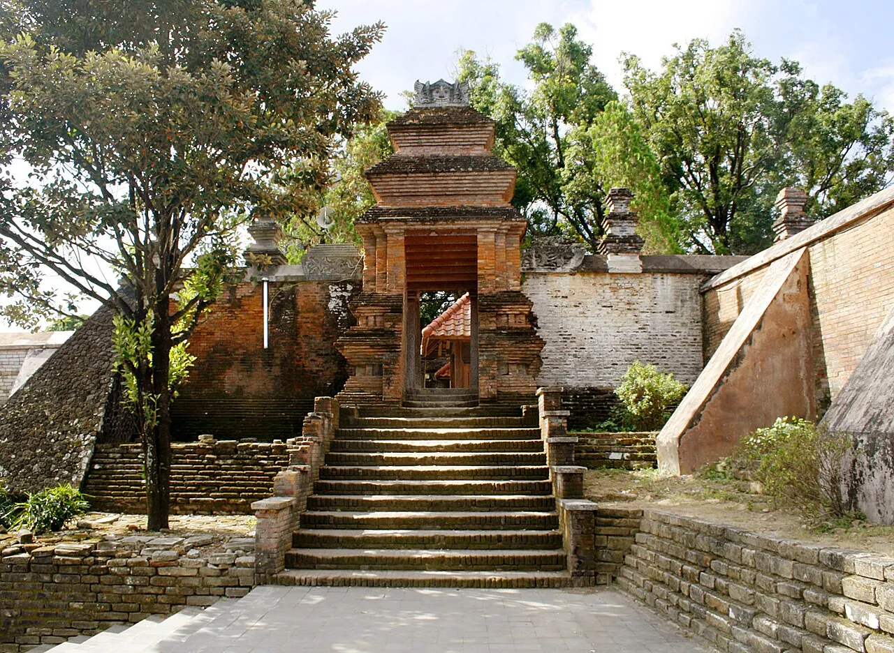 Portão de tijolo vermelho no alto de uma escadaria de pedra, entre muros antigos e árvores.