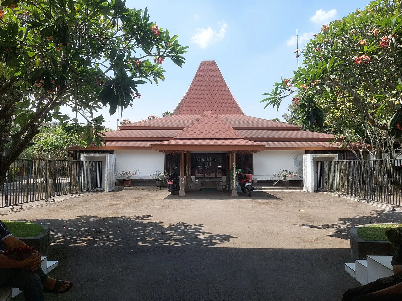 Edifício de paredes brancas e telhado vermelho alto em pirâmide, visto do pátio entre árvores floridas e um portão de grade.