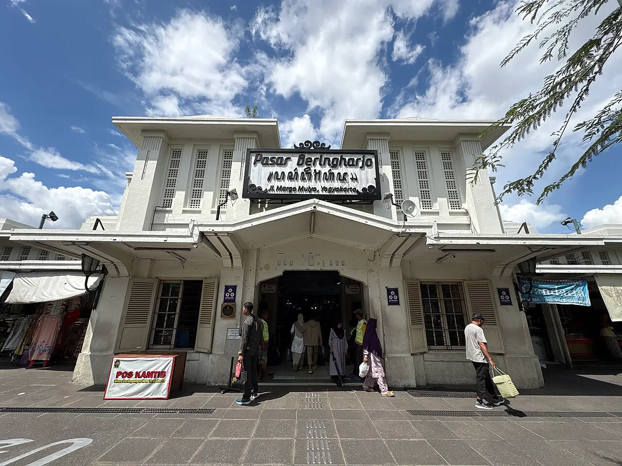 Fachada branca de um mercado com letreiro escuro sobre a entrada, pessoas entrando e saindo e céu azul com nuvens.
