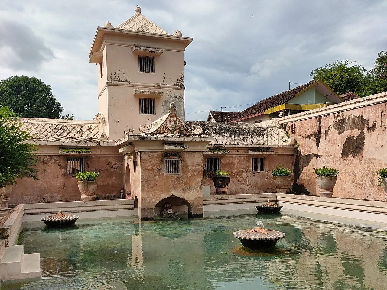 Piscina de água verde com fontes em forma de vaso, cercada de muros claros desgastados, vasos de plantas e uma torre de dois andares ao fundo.