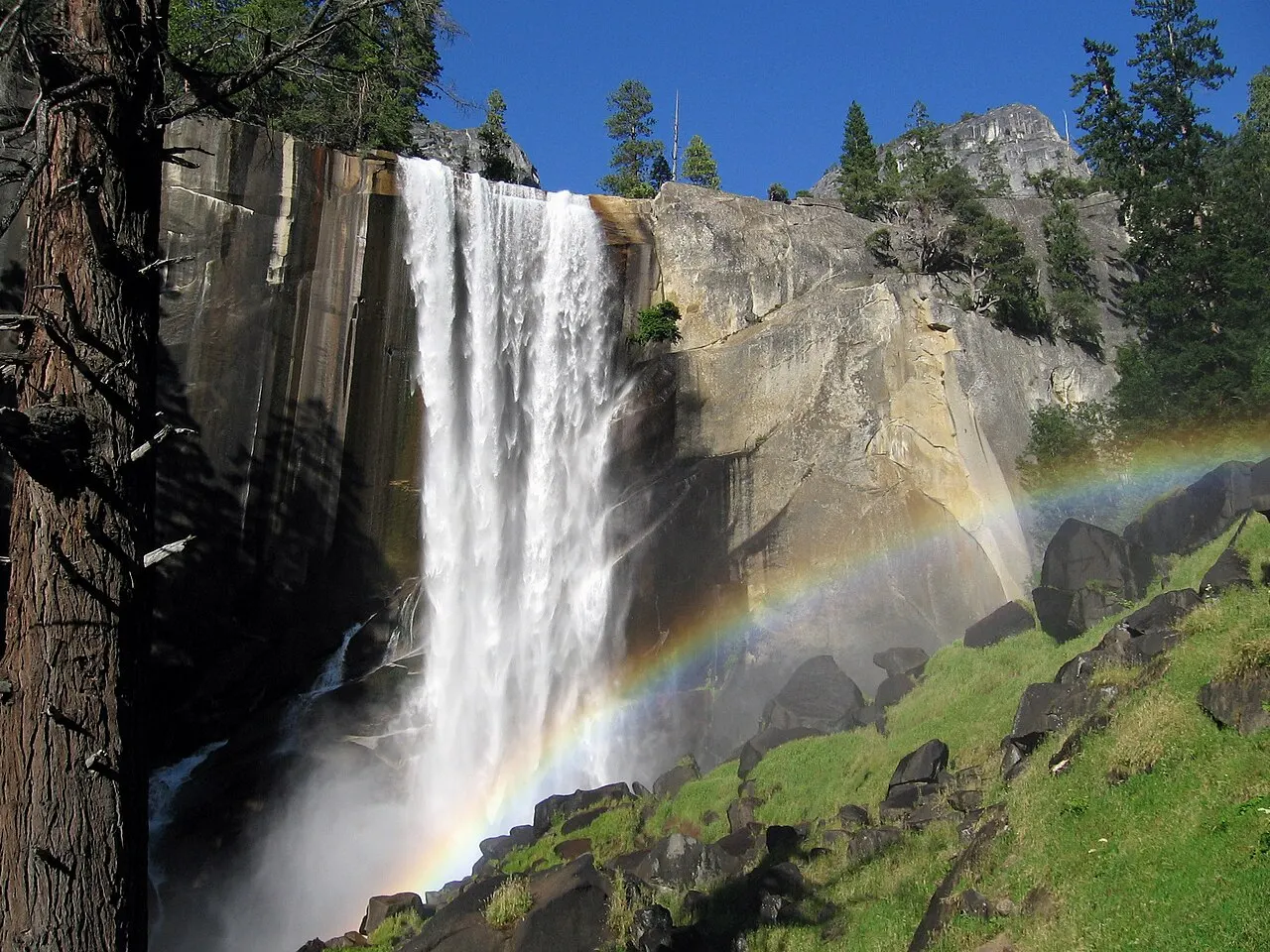 Uma cachoeira larga cai de um paredão de granito, com um arco-íris na névoa e rochas na encosta verde.