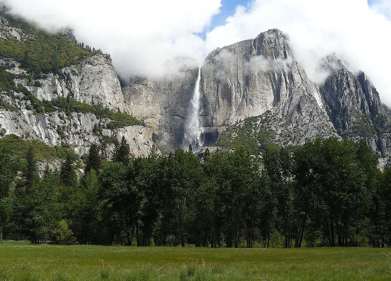 A cachoeira desce entre paredões de granito com nuvens no topo, vista de um prado verde com árvores na frente.