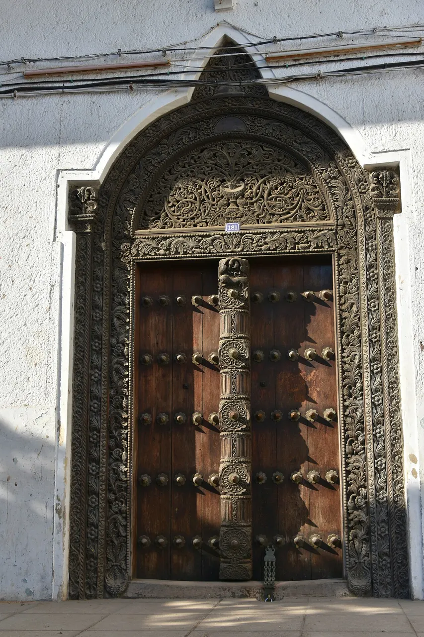 Porta de madeira esculpida, com cravos de metal e moldura trabalhada, numa parede branca de Stone Town.