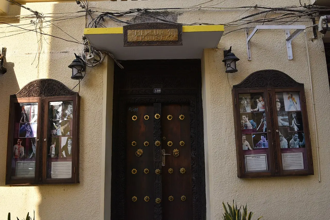 Porta da casa de Freddie Mercury, de madeira com rebites, entre duas vitrines de fotos na parede amarela.