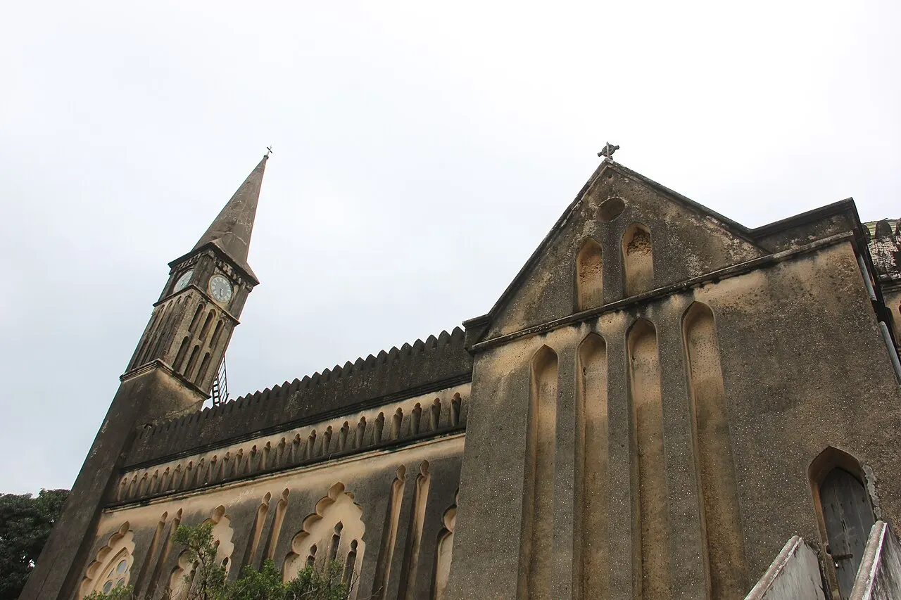 Torre esguia com relógio da Catedral Anglicana de Christ Church, ao lado do frontão em pedra com janelas em arco, sob céu claro.