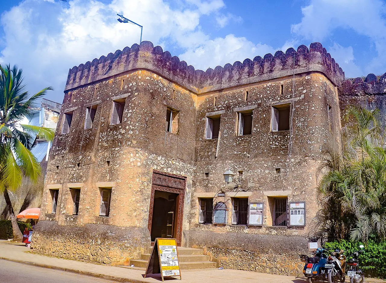 Entrada do Forte Velho de Zanzibar, um prédio de pedra coralina com ameias no alto e palmeiras ao lado.