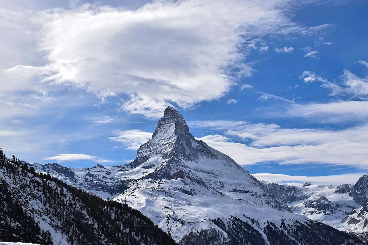 O pico em pirâmide do Matterhorn coberto de neve, com nuvens brancas no céu azul e encostas nevadas em volta.