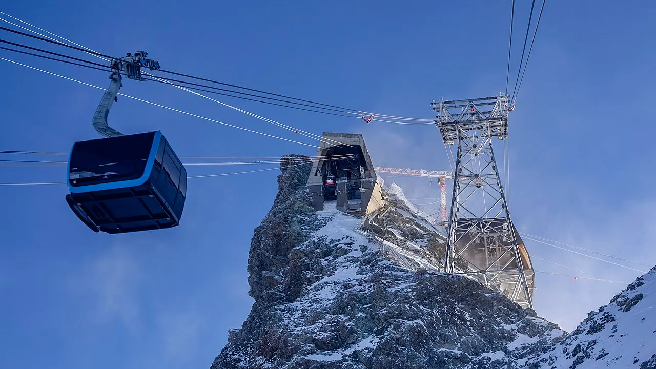 Uma gôndola do teleférico chegando à estação no pico rochoso do Klein Matterhorn, com a torre de aço e o céu azul.