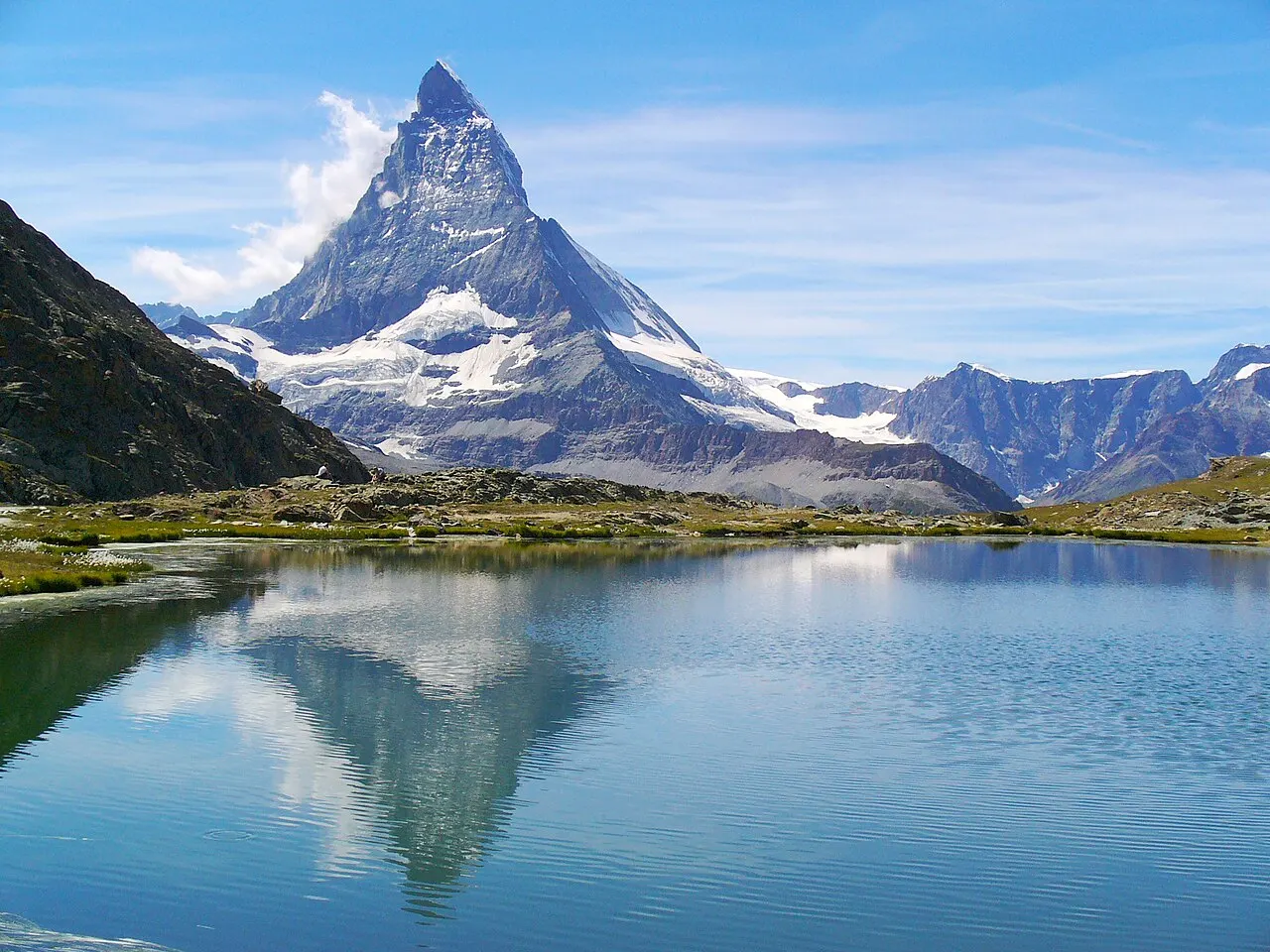 O lago Riffelsee com a água parada refletindo o Matterhorn nevado, sob céu azul.