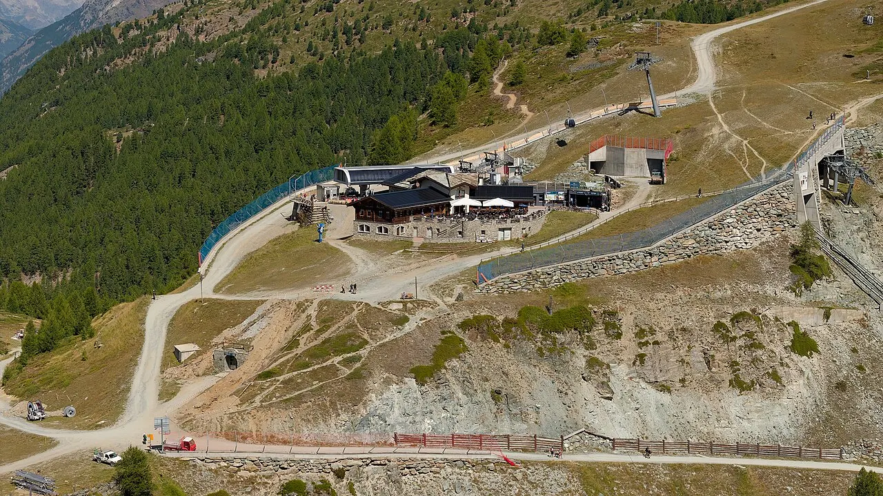 A estação e o restaurante de Sunnegga vistos do alto, na encosta seca, com trilhas, um teleférico e a floresta abaixo.