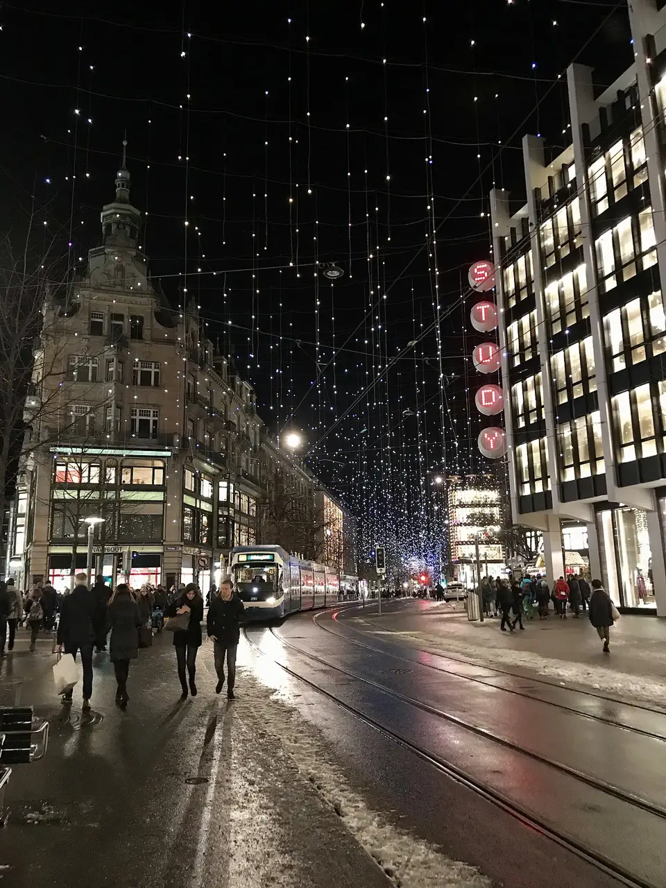 Bahnhofstrasse à noite, com o chão molhado, trilhos de bonde, luzes suspensas e pessoas nas calçadas.
