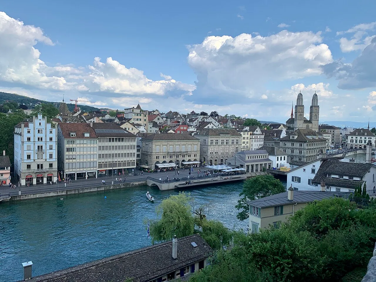 Vista do alto do Lindenhof sobre o rio Limmat, com prédios históricos na margem e as torres da igreja Grossmünster ao fundo, sob céu com nuvens.