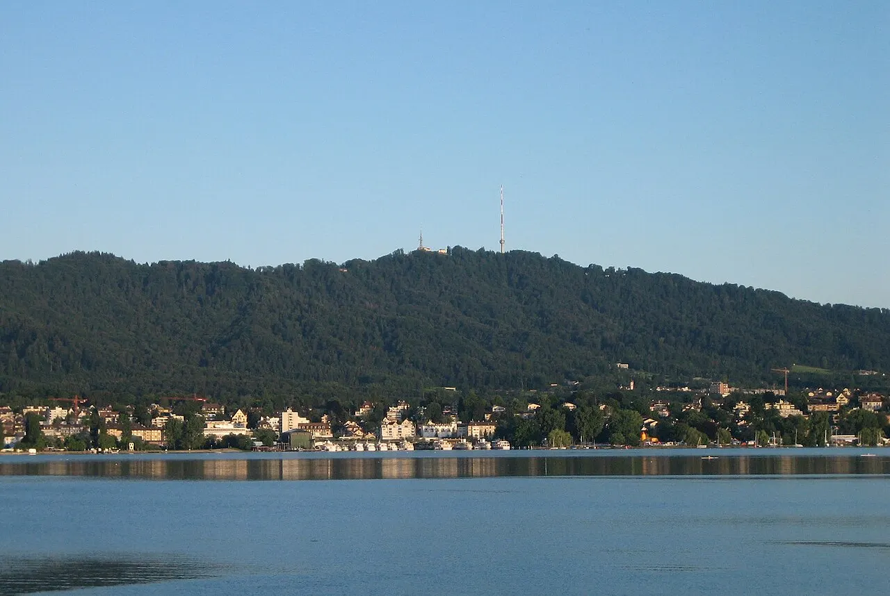 A colina arborizada do Uetliberg vista do lago de Zurique, com uma torre de transmissão branca na crista e casas baixas na margem.