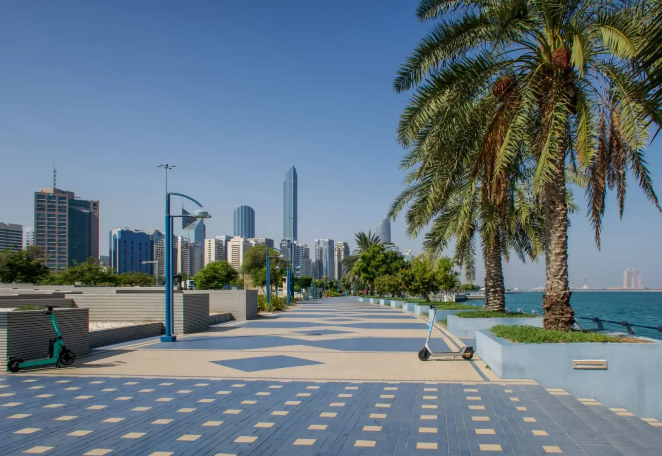 Passeio da Corniche em Abu Dhabi, com palmeiras, patinetes, edifícios ao fundo e o mar à direita.