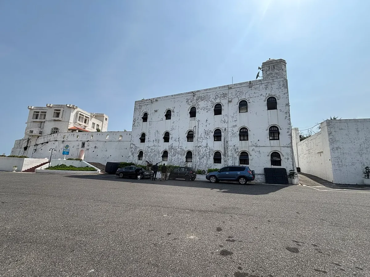 Pátio do Castelo de Osu em Acra, com fachadas brancas, torre e hastes no telhado, carros e visitantes junto ao edifício e pavimento frontal.