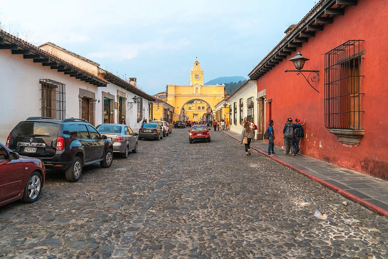 Arco de Santa Catalina com relógio e cruz, visto pela rua de Antigua; casas, luminárias, carros e visitantes nas laterais, montanha e céu ao fundo e pavimento de pedras à frente.