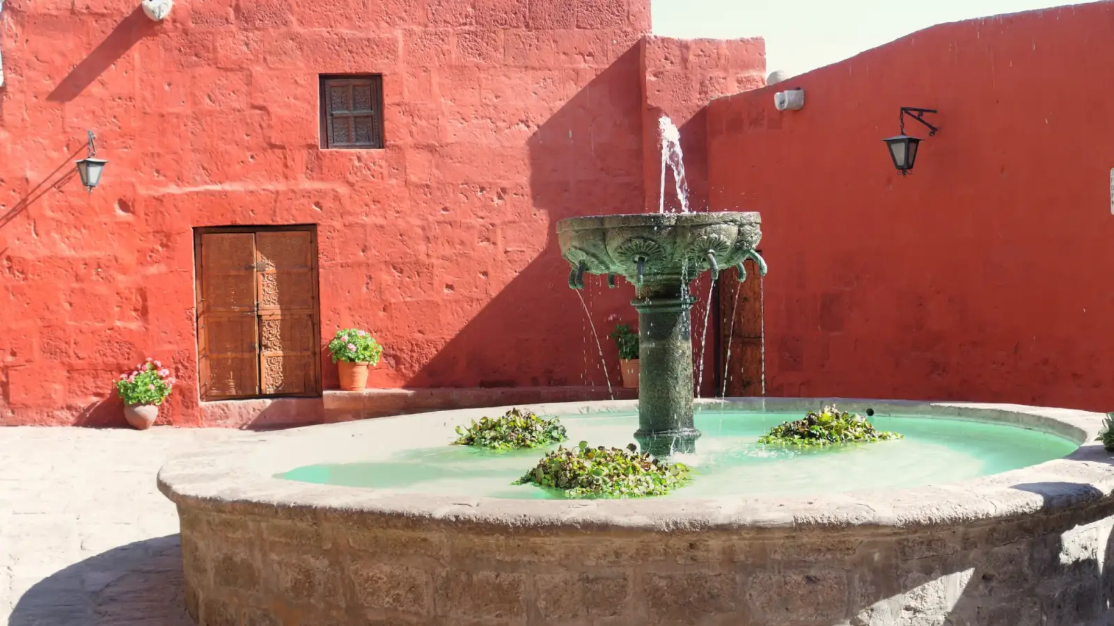 Um pátio do Monasterio de Santa Catalina com muros pintados de vermelho, porta de madeira, vasos de flores e um chafariz de pedra com água turquesa no centro.