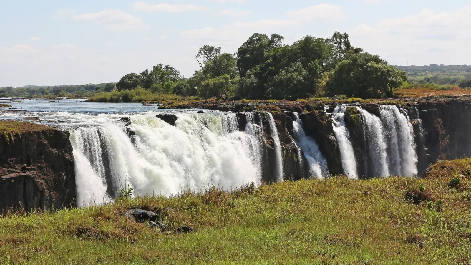 Les chutes Victoria vues du côté du Zimbabwe : l'eau blanche tombe en plusieurs rideaux par le rebord de roche sombre, avec des arbres au-dessus de la falaise et de l'herbe sur la rive au premier plan