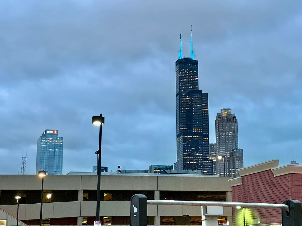 A Willis Tower e suas duas antenas iluminadas, com outros prédios do centro atrás de uma garagem e postes sob céu nublado.