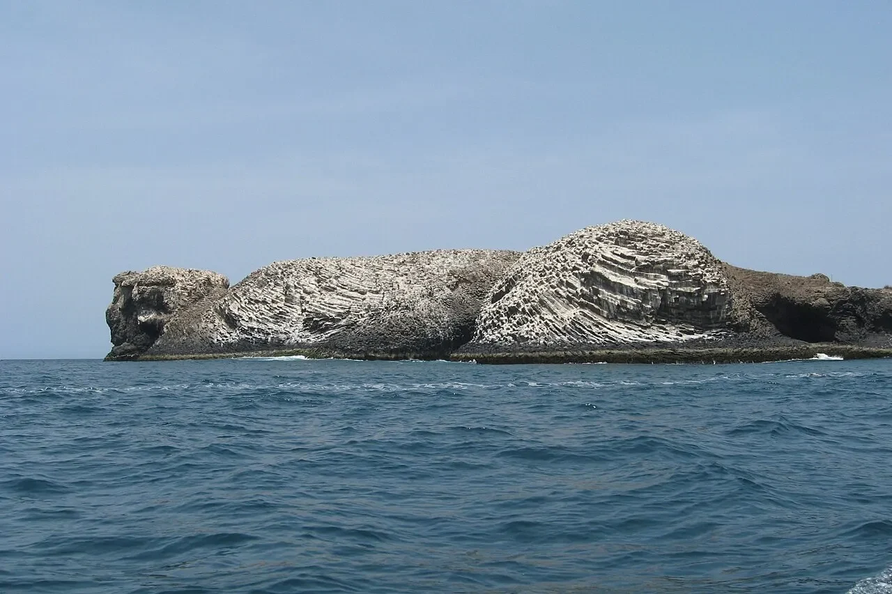 Rochedo estratificado das Îles de la Madeleine ao largo de Dakar, com céu azul ao fundo, costa escura e ondas do Atlântico em primeiro plano.