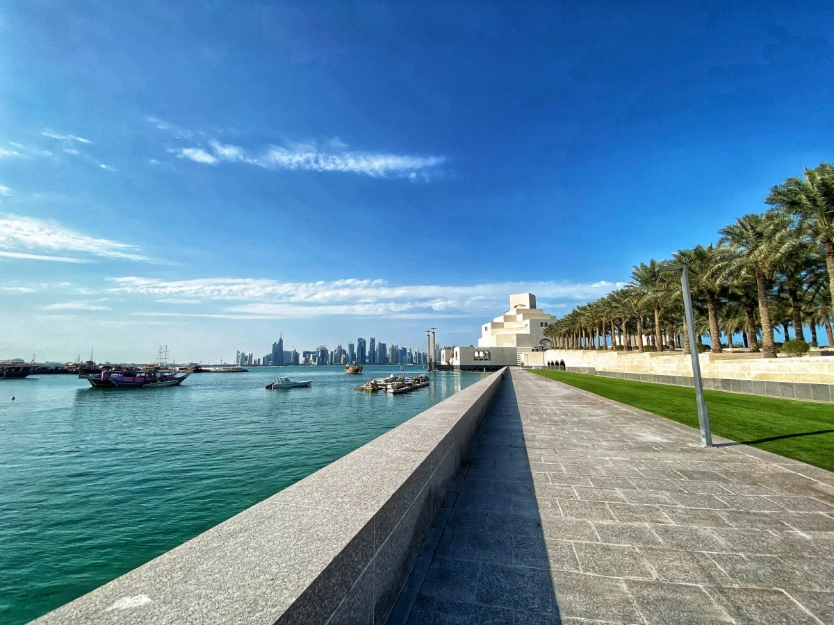 Passeio do MIA Park à beira da baía, com muro de pedra e luminária em primeiro plano, Museu de Arte Islâmica e palmeiras à direita e barcos diante do skyline de Doha.