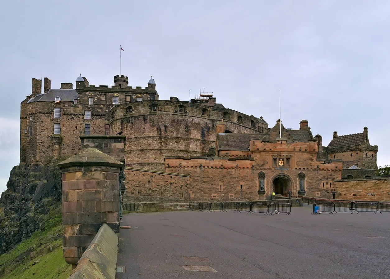 Castelo de Edimburgo visto da esplanada, com bandeira, mastros, muralhas e portal; pilar e balaustrada de pedra à frente, céu nublado e pavimento com barreiras temporárias.