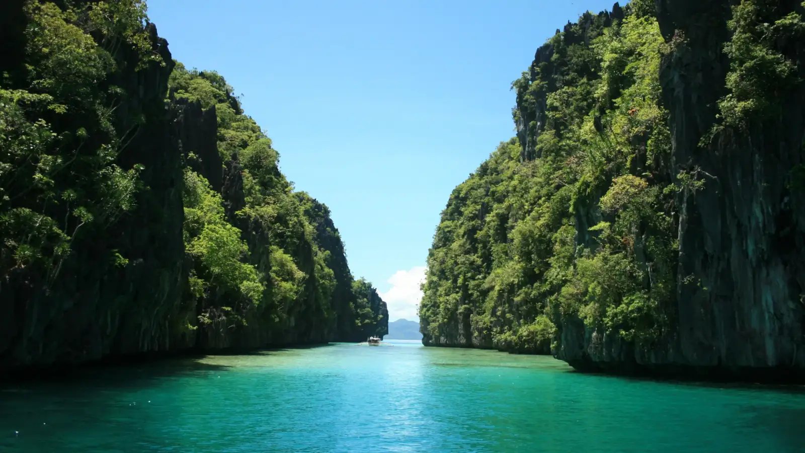 The entrance to the Big Lagoon in El Nido: a channel of turquoise water between two limestone cliffs covered in forest, with a small boat in the distance and a blue sky.