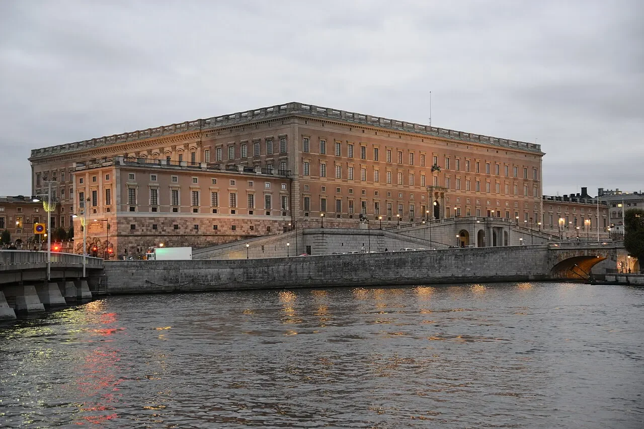 Palácio Real de Estocolmo ao entardecer, com mastro no telhado, fachadas de pedra, rampas, muro do cais e ponte; água com ondas e reflexos em primeiro plano.