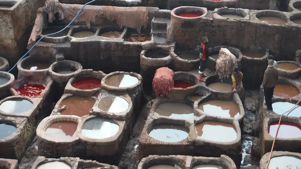 Vista de cima de dezenas de tanques redondos e retangulares de pedra, cheios de líquidos de cores diferentes como branco, vermelho e marrom, com peles penduradas e alguns trabalhadores caminhando entre os tanques.