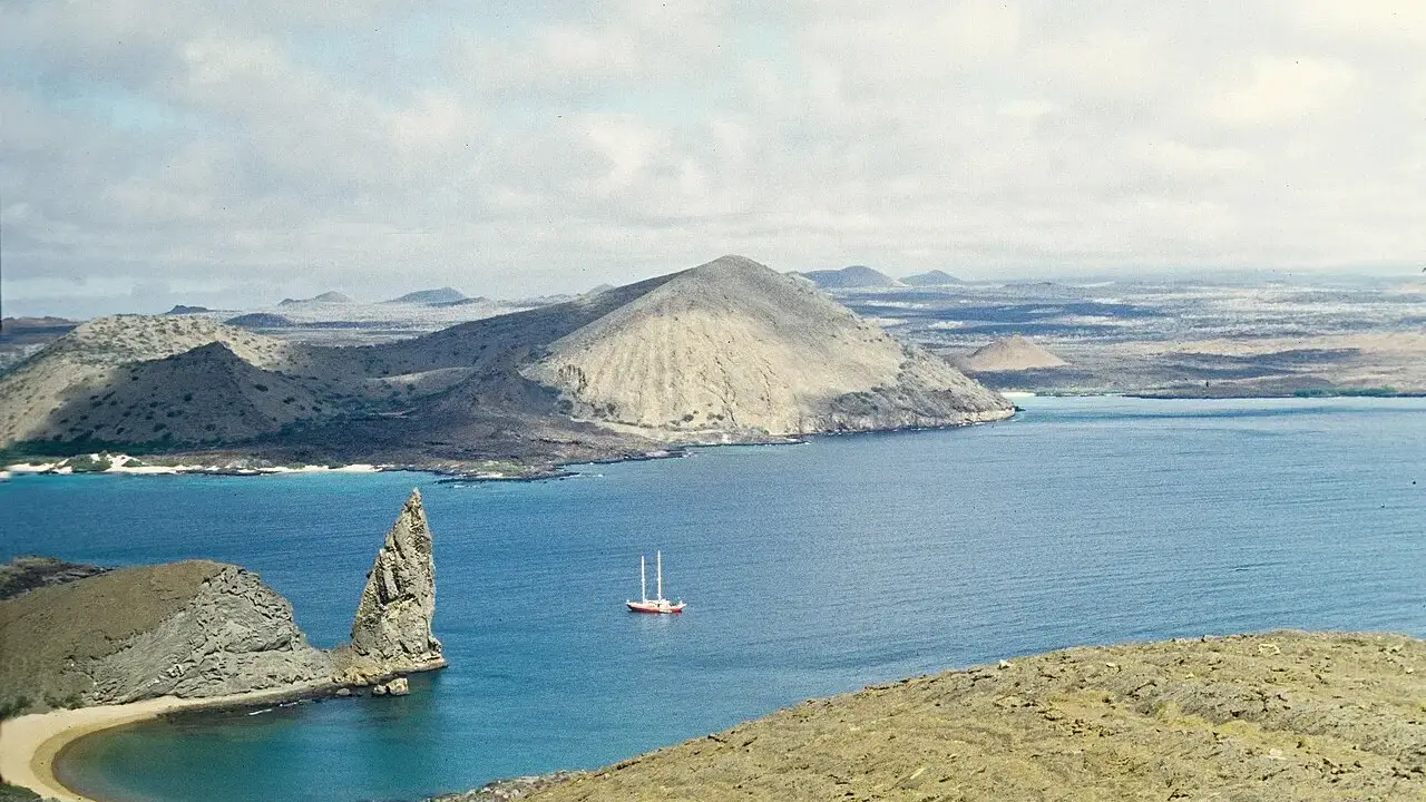 O obelisco de rocha do Pinnacle Rock na ponta de Bartolomé, com uma praia em meia-lua de areia clara à esquerda, um veleiro fundeado na baía azul e cones vulcânicos castanhos ao fundo.