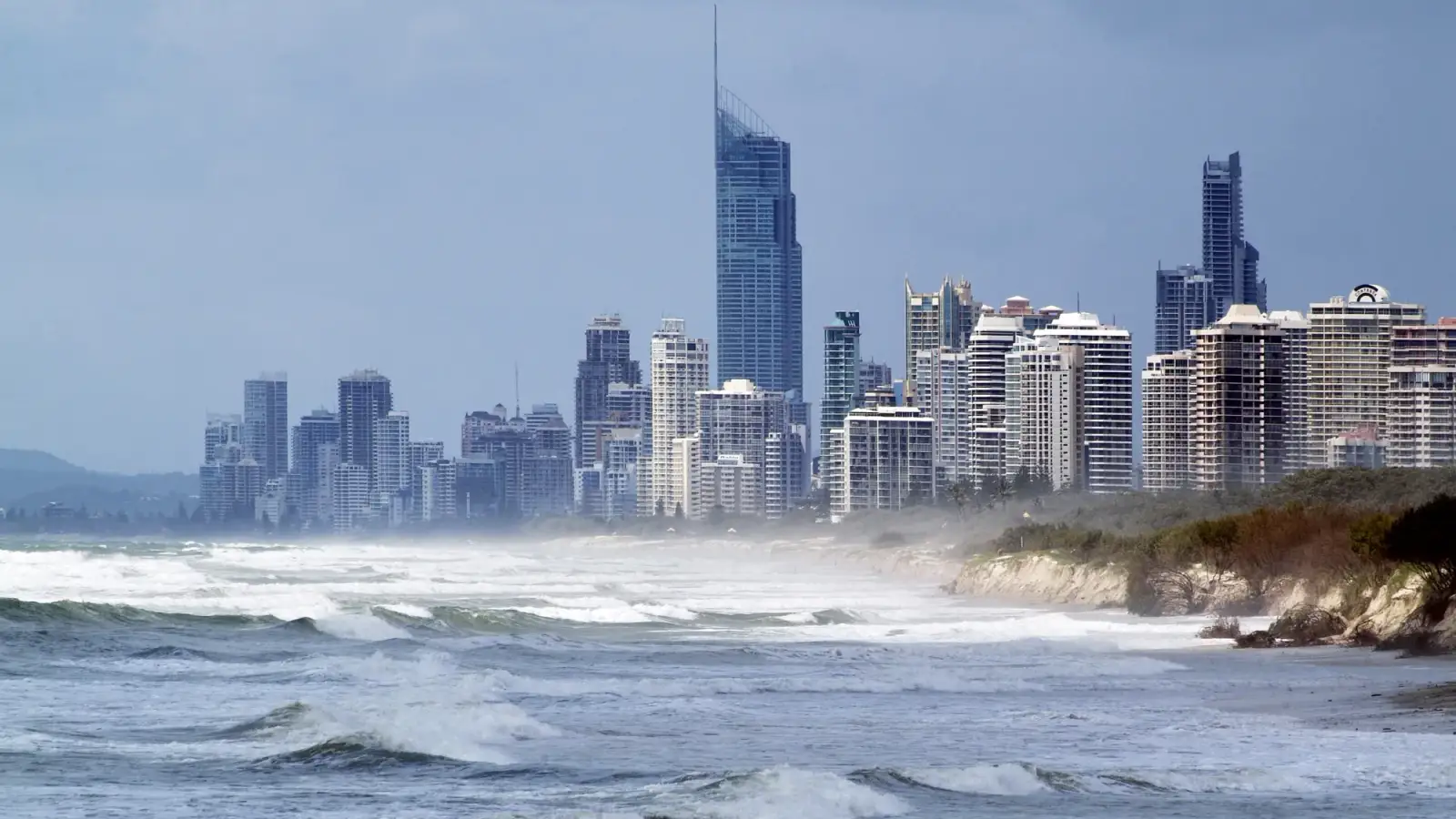 The high-rises of Surfers Paradise, with the Q1 tower rising in the middle, seen from the beach with breaking waves and a vegetated dune on the right, under an overcast sky