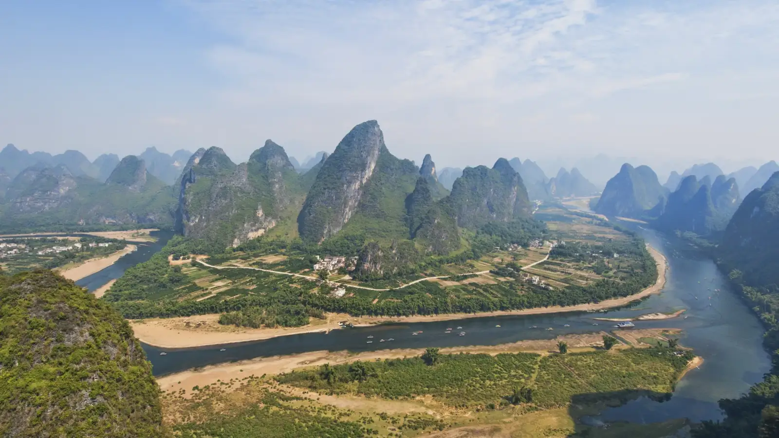 The Li River seen from above, curving between fields and sandbanks, with pointed forested limestone hills in rows to the hazy horizon