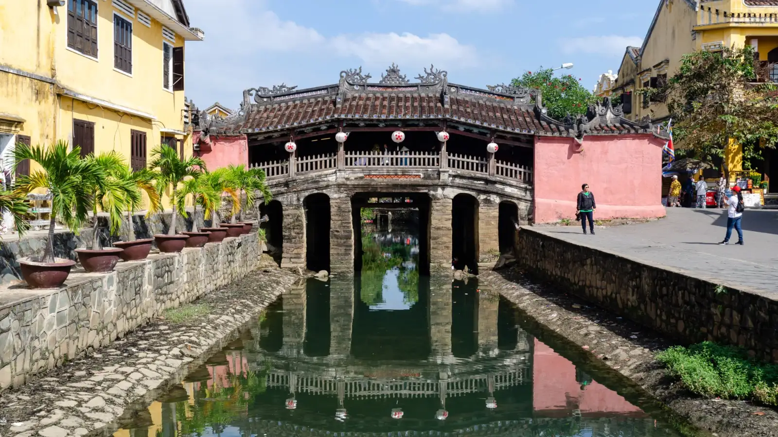 Le pont couvert japonais de Hội An, au toit orné et aux lanternes rouges, reflété dans le canal entre des maisons jaunes et des palmiers en pot