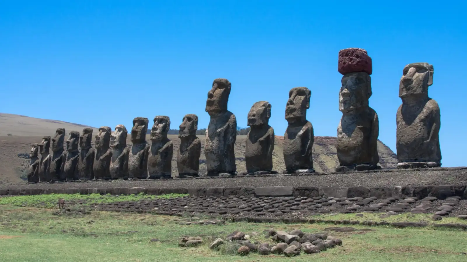 A fileira de quinze moais de Ahu Tongariki sobre a plataforma de pedra, um deles com o pukao vermelho, sob céu azul e com a encosta do vulcão atrás.