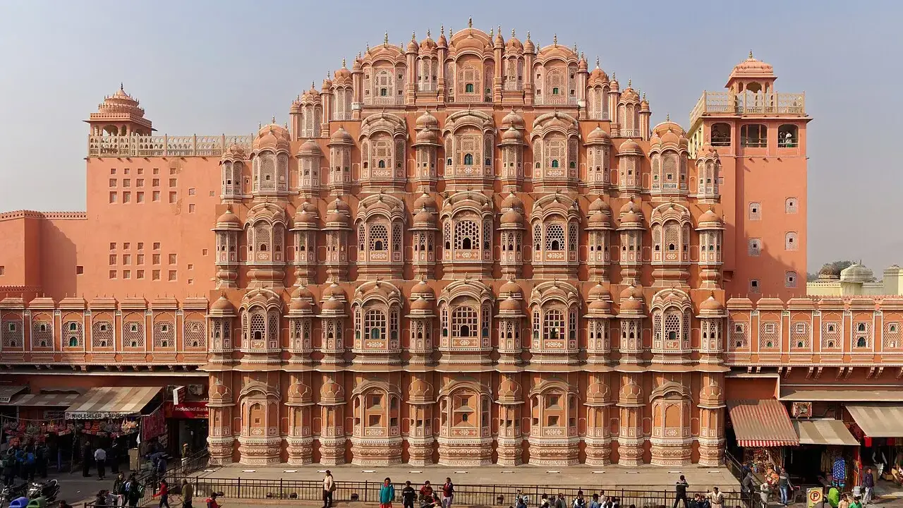 Fachada rosa inteira do Hawa Mahal, com as alas laterais e a rua com carros e pessoas na frente.
