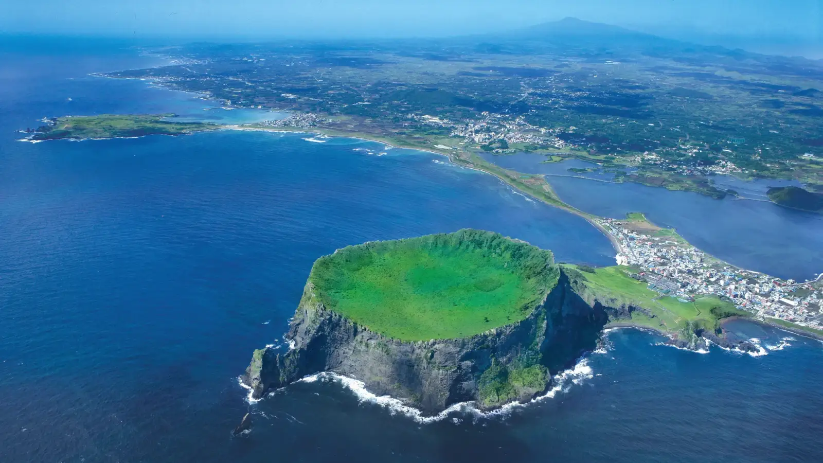El Seongsan Ilchulbong visto desde el aire, un cráter verde rodeado de acantilados sobre el mar, con el pueblo de Seongsan y el Hallasan al fondo