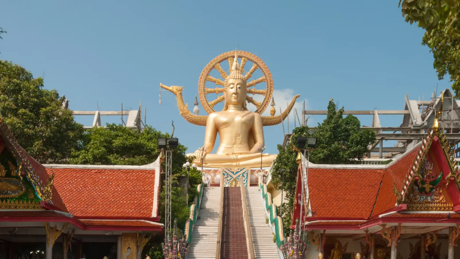 Le Big Buddha doré du Wat Phra Yai, à Koh Samui, assis devant une roue dorée en haut d'un escalier, entre deux pavillons aux toits rouges, des arbres et un ciel bleu.