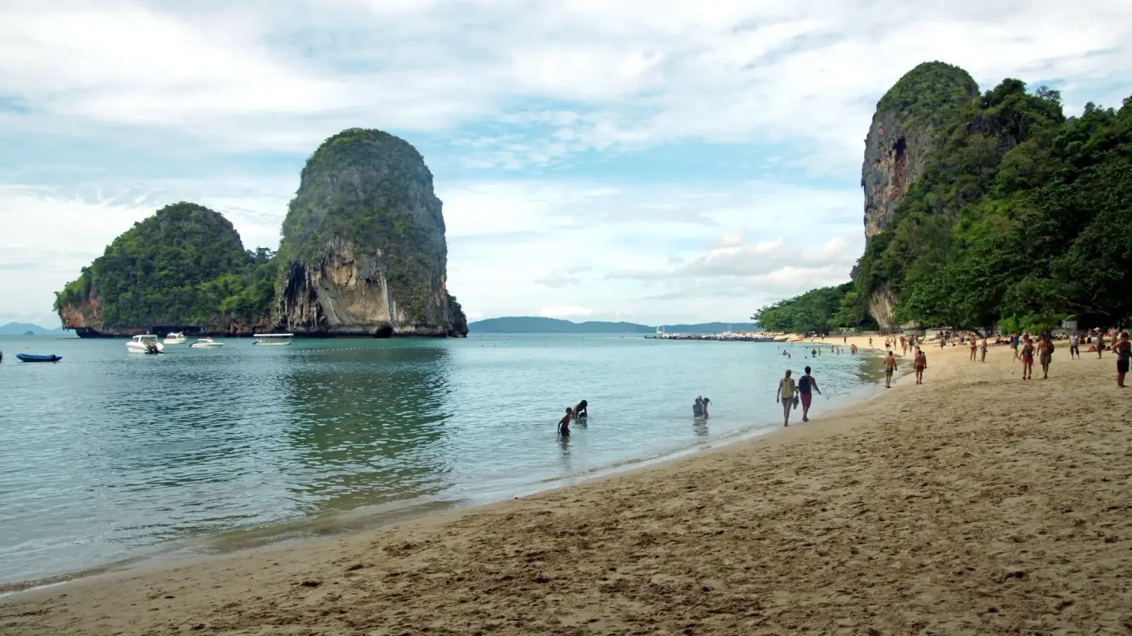 La plage de Phra Nang, à Krabi, avec l'îlot calcaire couvert de forêt dans la mer calme, des bateaux au loin et la falaise boisée à droite du sable